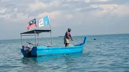 A small-scale fisherwoman stands in a small blue boat with a net flowing into the water.