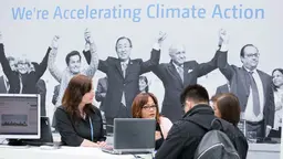 Two people taking to representatives at the Paris Agreement stand. Behind them on the wall, there is a big image of people holding hands with the words 'We're accelerating climate action'.