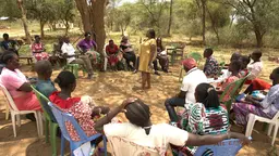 Community members sitting on chairs in a circle, with a person standing in the meeting talking and presenting.