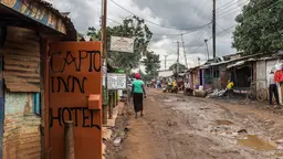 People walk on a muddy path between metal huts on either side of the 'road'. In the foreground a door to a building is open and written on it is 'Capto Inn Hotel'. 