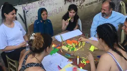 People sitting around a table, discussing and building model houses out of colorful paper materials and glue.