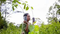 A woman farmer in a field carries a small plant in each hand at shoulder height.