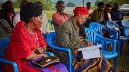 Group of community members Kibaale, Uganda are sitting down and discussing during consultations with officials. Two women are sitting on blue chairs, listening and noting things down on notebooks.