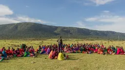 Group of women and young girls dressed in traditional clothing, sitting down in a wide open field, in a circle. Another person is standing and talking to them.