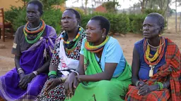 Four women seated in a line and wearing colourful traditional pastoralist clothing lean forward as part of an interview being conducted outside, with trees in the background.