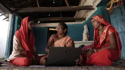 Three women wearing tradition sarees sitting down and discussing. One of them is holding a laptop and explaining something to the other two women.
