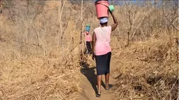 A woman balancing a bucket of water on her head in pictured from behind as she walks through a field. 