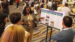 Person standing next to a printed out poster, pointing and speaking. Other people are gathered and listening.