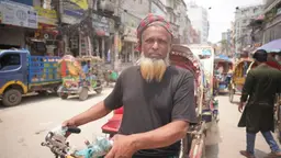Older man standing in a middle of a busy street in Dhaka, looking straight at the camera. He is wearing a grey t-shirt and a red scarf on his head, and holding his motorcycle.