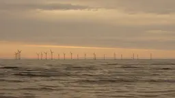 A dramatic sunset of reds and yellows frames a dozen wind turbines in the distance in the water.