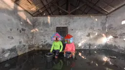 Two people wearing boxes painted like houses on their heads sit in knee-deep water inside a derelict house.