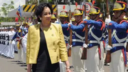 A woman in a bright yellow jacket stands next to parading troops.