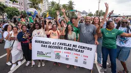 A group of men and women, cheering and chanting and waving their hand in the air, pose behind a sign in Brazilian Portuguese that translates as "We want the favelas to be at the heart of climate decisions".