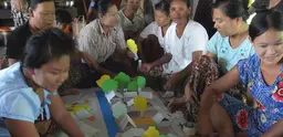 Women from near Rangoon, Myanmar are seated around a map with paper houses and trees talking and laughing.