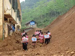 Children with backpacks walking along a path with a building on the left and a forest and looming hills behind.