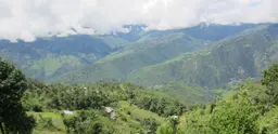 Forested hills in Nepal, with a house in the foreground and forested hills, and then clouds behind.