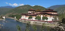 Picture of the Punakha Dzong, an administrative centre in Bhutan. A river runs past it in the foreground and wooded hills rise behind it.  