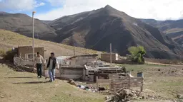 Two men stand in front of a building under construction, with mountains in the background