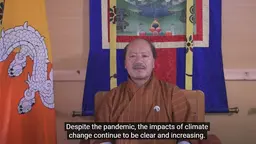 A seated man gives a speech to a camera directly in front of him, next to the Bhutan national flag