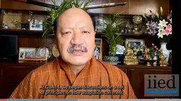 Head and shoulders image of Sonam P Wangdi, in traditional Bhutanese attire, speaking into a video camera 