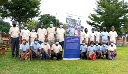 A group of people pose for a photo outside, with trees in the background. There is a banner in the middle