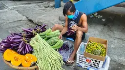 Man sits on the floor holding aubergines. In front, there's a table with vegetables
