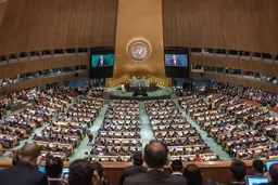 A large audience sits in a cricualr room, facing a man behind a lectern. The UN logo is printed on the wall.