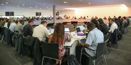 A large number of people, with backs to the camera, are seated around a table. On the tables there are cards indicating which countries they represent