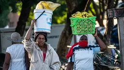 Two women carry buckets with fruits on their heads.