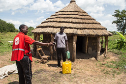 Man stands next to a hut and a jerrycan. Another man, wearing red, points to the right.