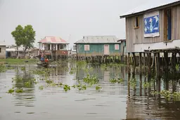 Two people sail a canoe around stilt houses.