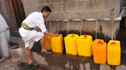 A man lines up six bright water bottles under a series of outside taps