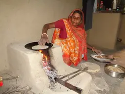 A woman crouches on the flor next to a stove to make a meal