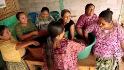 A group of women laugh and smile as they stand and dip their hands into two large buckets on a table 