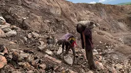 Two men on the side of a mountain; one bends over to pick up two sacks, the other carries them on his shoulders