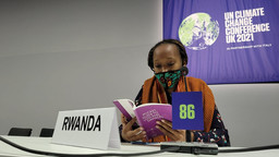 A masked woman sits behind a desk reading an information booklet next to a sign that is labelled 'Rwanda'. behind her is a large banner proclaiming the 2021 UN Climate Change Conference