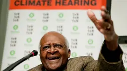 Close up of a man speaking to a microphone and his hand raised. Behind him a sign says "climate hearing".