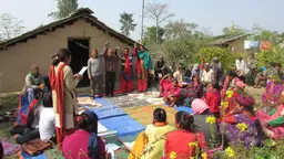 A group of people sit on the floor in a circle. A woman on her feet is reading from a paper. Some other people are no their feet and holding hands in the air.