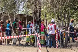 Women stand in a line with empty bags. A man wears a white vest with a red cross on the back.