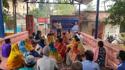 People sit on the floor in  group under cover facing a standing man making a presentation/speech