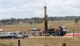 Fenced working site in a field. There are workers and cars parked and a mineral exploratoion drilling rig