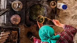 Zenital view of a woman, wearing a headscarf, holding a spoon and a bowl with greens in front of a pot with more greens.