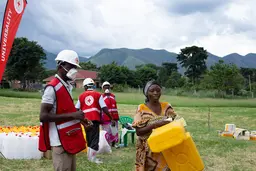 A woman carries jerrycans and tree men behind her wear masks and red vests with a red cross in the back