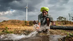 A man crouches by a pipe as water erupts from it through his cupped hands on to his field, with a wind turbine in the background.