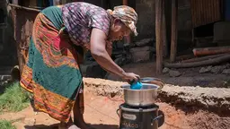 Woman bends over to put a small plastic cup in a cooking pot, which is on a stove.