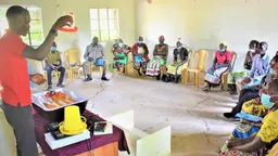 A man stands behind a desk raising an object in the air as he makes a demonstration to people seated in a semi-circle around him.