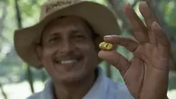 Man smiling broadly holds up to the camera a fragment of gold between his thumb and finger