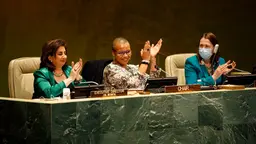 Three women, one wearing a mask, the other two smiling and clapping, sit at a raised desk in front of an audience