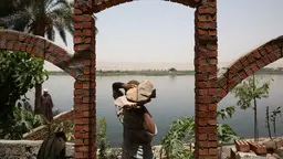 A man, his head hidden by the two large rocks he carries on his shoulders, on the edge of a large river 