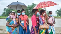 A group of woman wearing bright clothes and masks huddle under umbrellas, with a flooded river behind. 